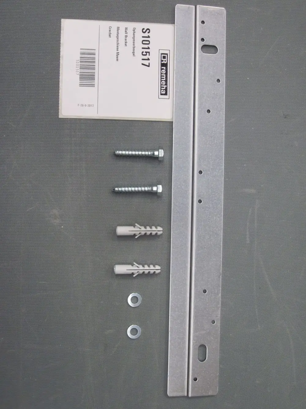 Montageschiene von Remeha, begleitet von zwei Schrauben, zwei Dübel und zwei Unterlegscheiben, arrangiert auf einer grauen Unterlage.